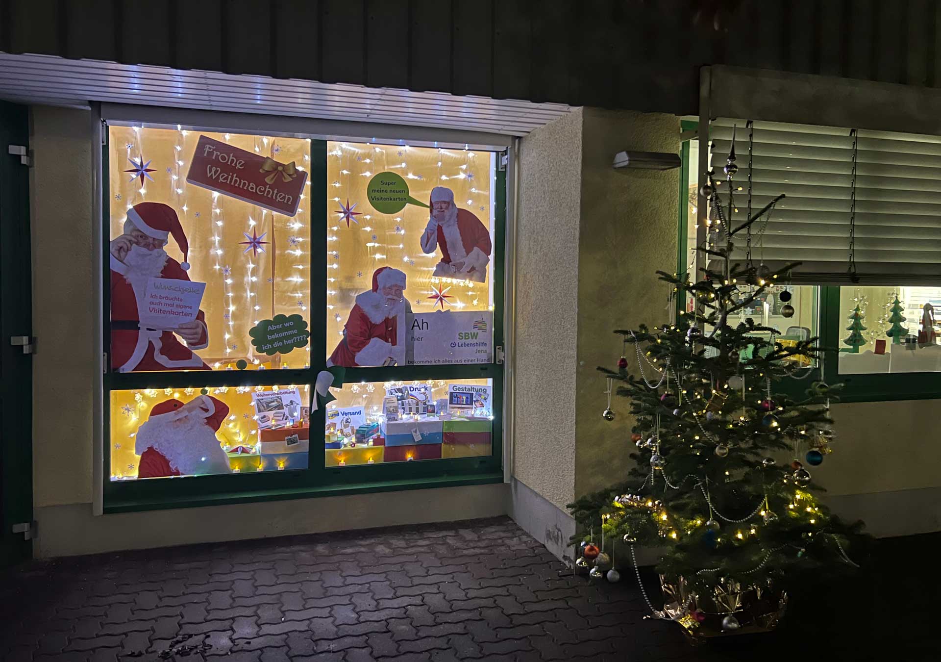 Das Bild hat eine weihnachtliche Abendstimmung. Rechts ist ein geschmückter Weihnachtsbaum, links ein geschnücktes Fenster mit Lichtern und Weihnachtsmännern, die sich eine Visitenkarte drucken wollen.