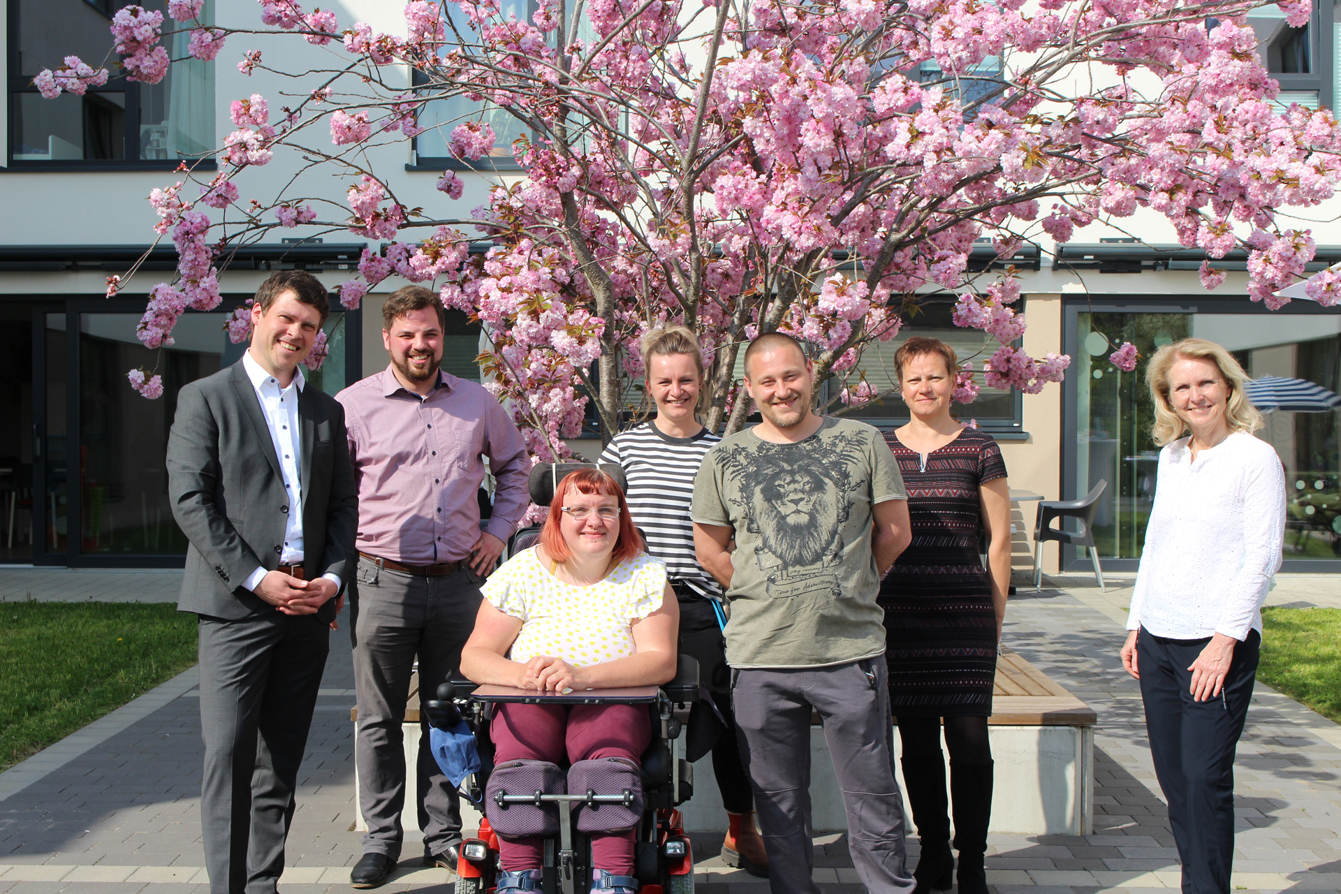Eine freundliche Gruppe von Menschen mit und ohne Behinderung steht vor einem blühenden Kirschbaum vor der Wohnanlage Gartenhof.