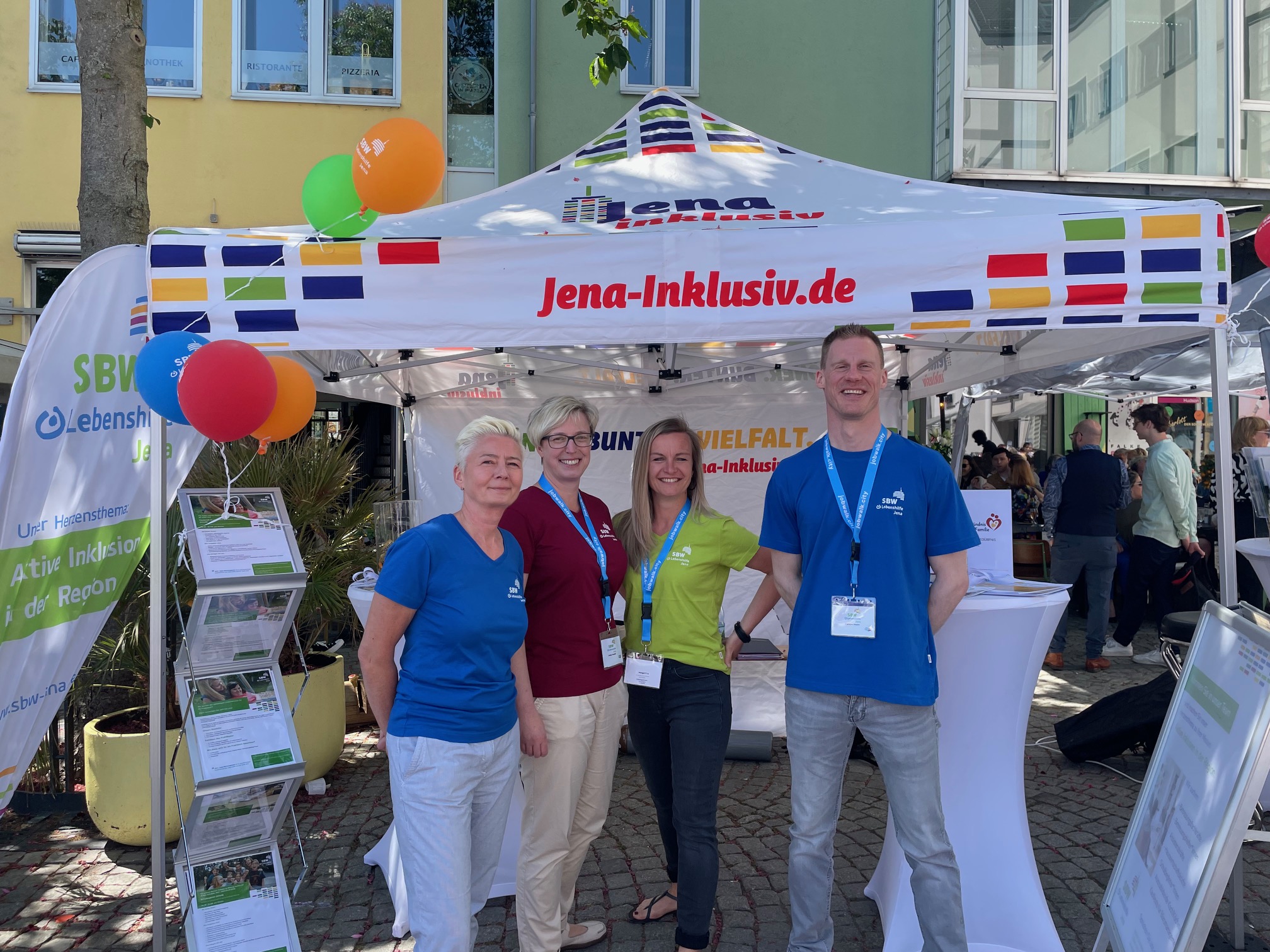 Vier Personen in farbigen T-Shirts stehen freundlich lächelnd vor einem Pavillion mit der Aufschrift Jena-Inklusiv