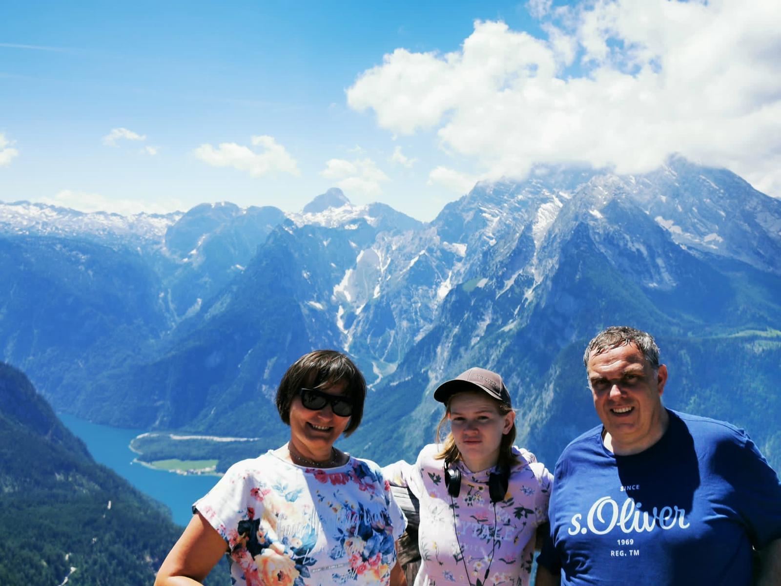 Das Bild zeigt drei Personen vor einem Bergpanorama. Auf den Gipfeln sieht man noch Schneereste und im Tal einen blauen See.