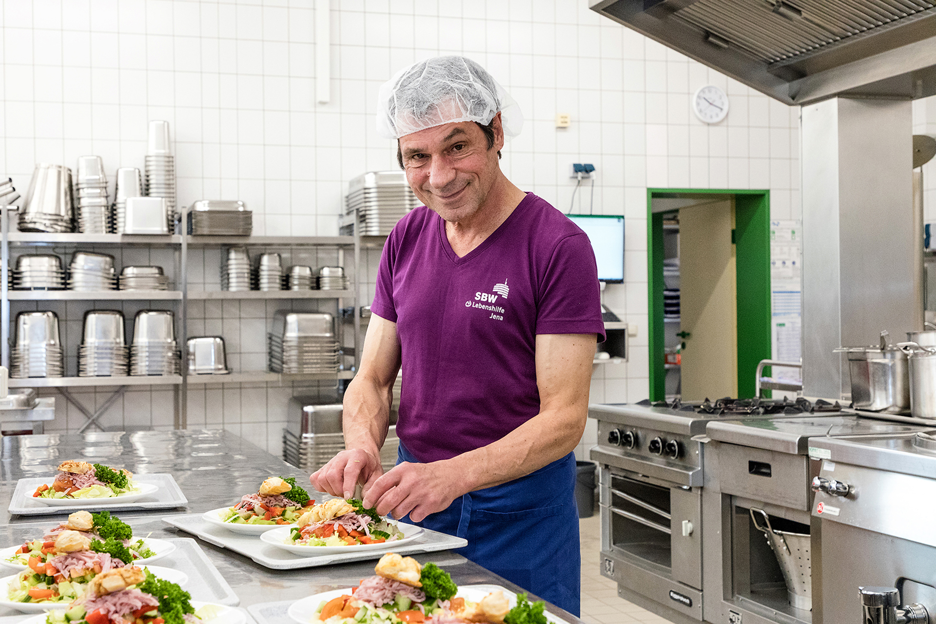 Ein Mitarbeiter bereitet Salatteller zu. Im Hintergrund sieht man die Ausstattung der Küche.