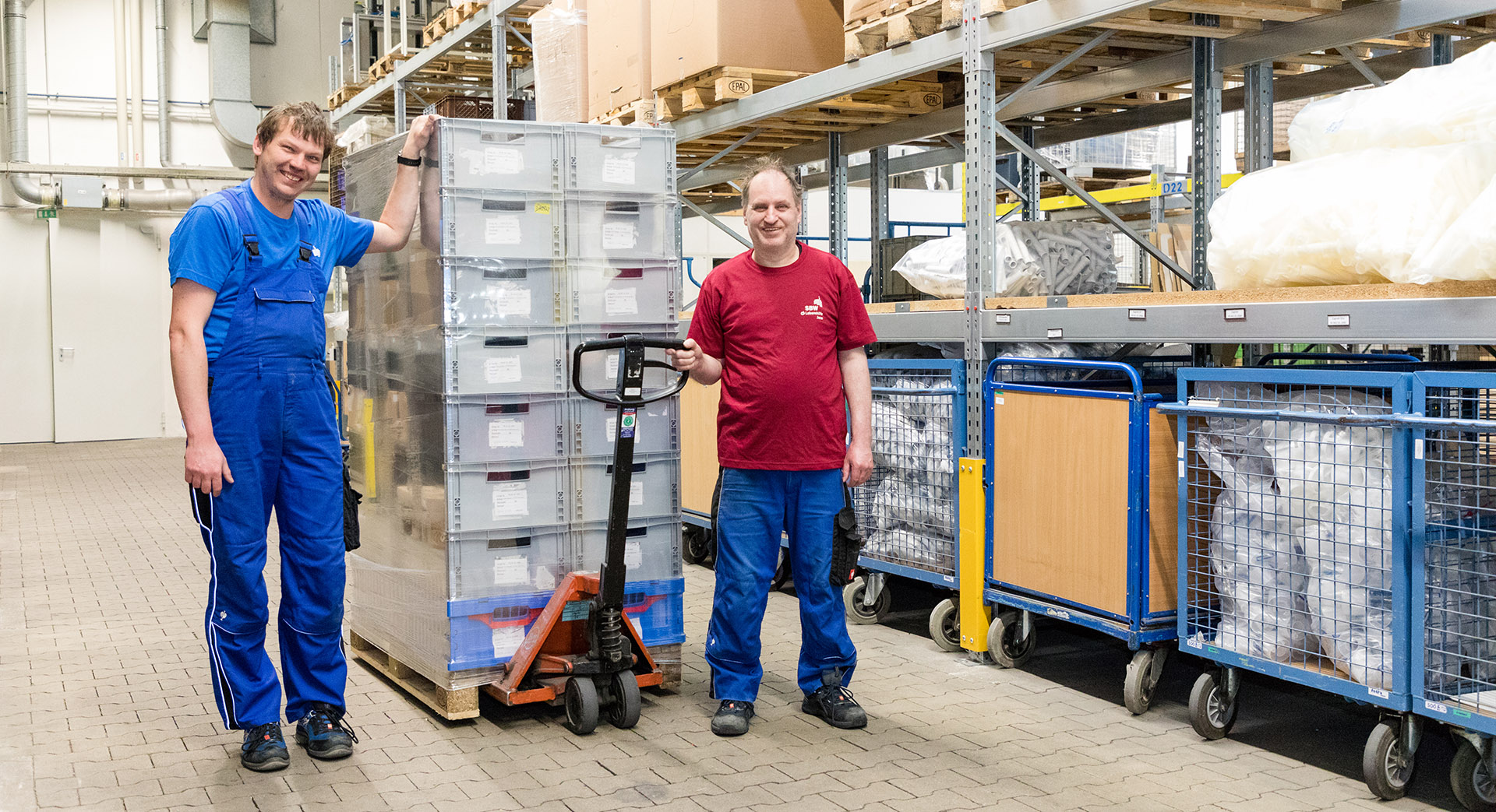 Zwei Mitarbeiter stehen an einem Hubwagen. Auf dem Hubwagen ist eine Palette mit Kisten.