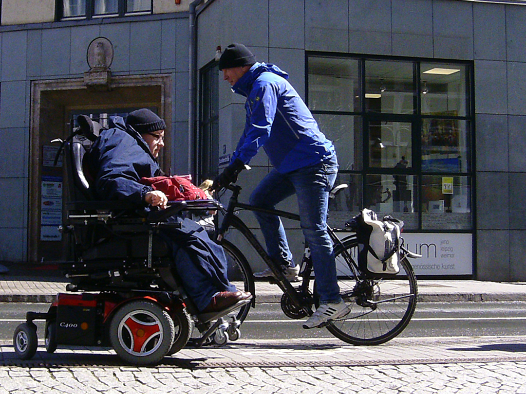 Im Bild zu sehen ist eine Frau im Rollstuhl in einer Straßen-Szene.