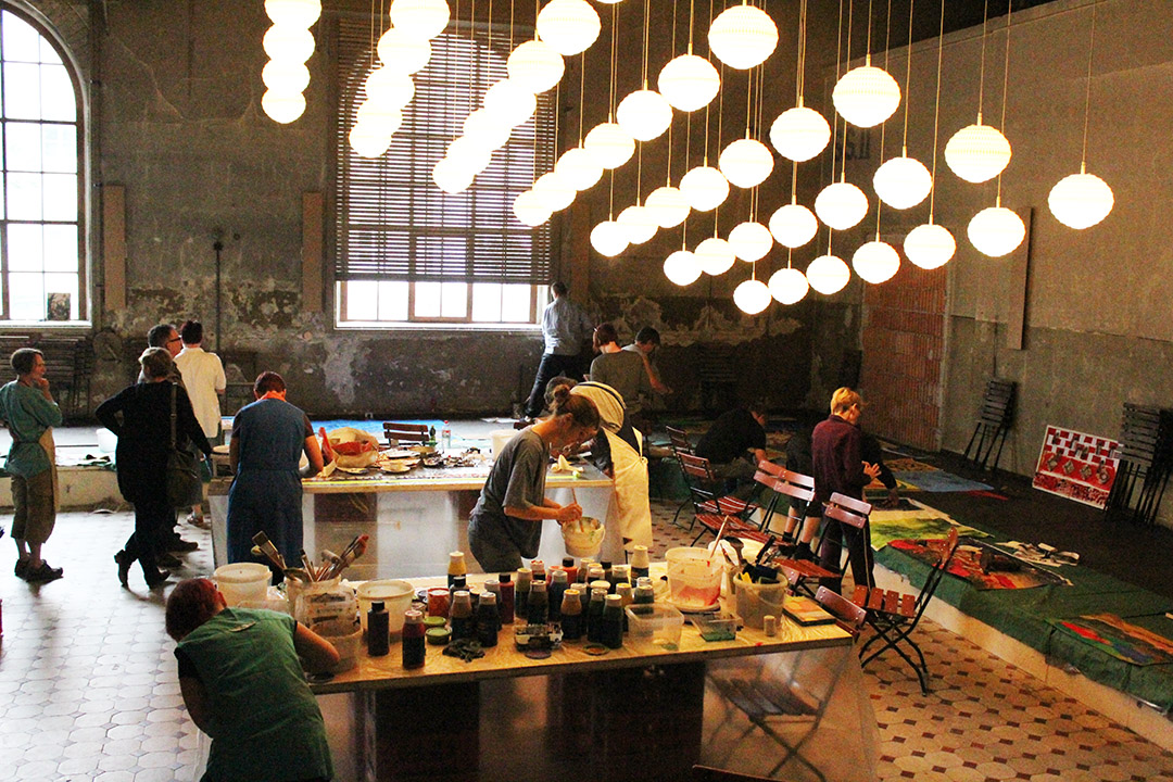 Im Bild zu sehen sind Personen in einem alten Industriegebäude. Auf den Tischen stehen viele Farben und auf dem Boden sieht man Kunstwerke.