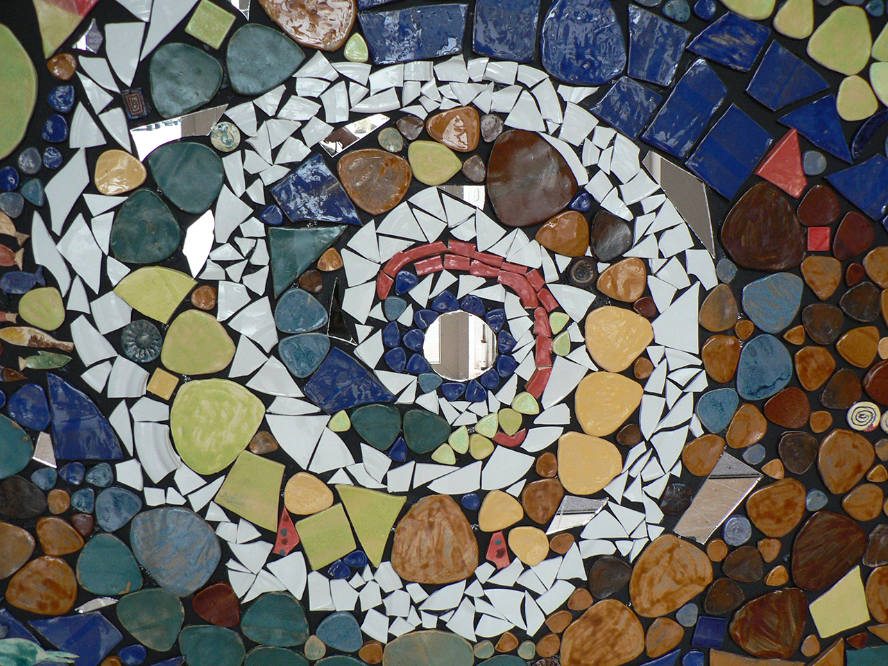 Im Bild zu sehen ist eine kunstvolle Mosaik-Spirale. Sie besteht aus Keramik-Teilen in weiß, blau, braun und rot sowie grün.