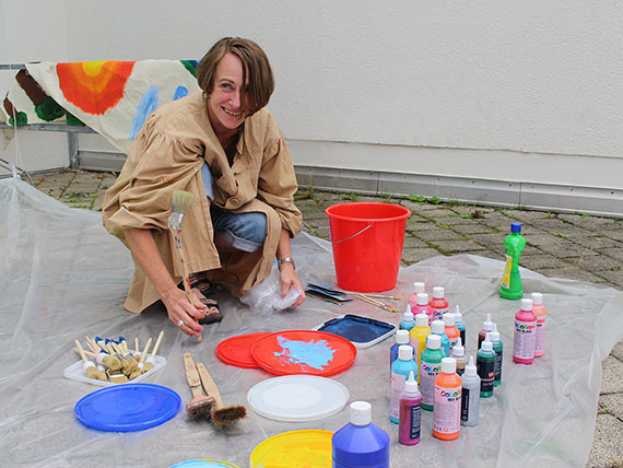 Zu sehen ist die Projekt-Koorinatorin Christine Klemm. Sie hockt auf einer Plastik-Plane umgeben von bunten Farb-Behältern und Pinseln.