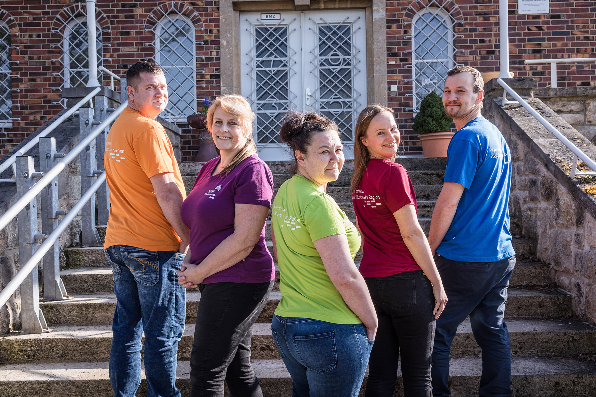 Ein Mitarbeiter-Team steht auf einer Treppe. Alle tragen bunte SBW-Shirts und schauen freundlich.