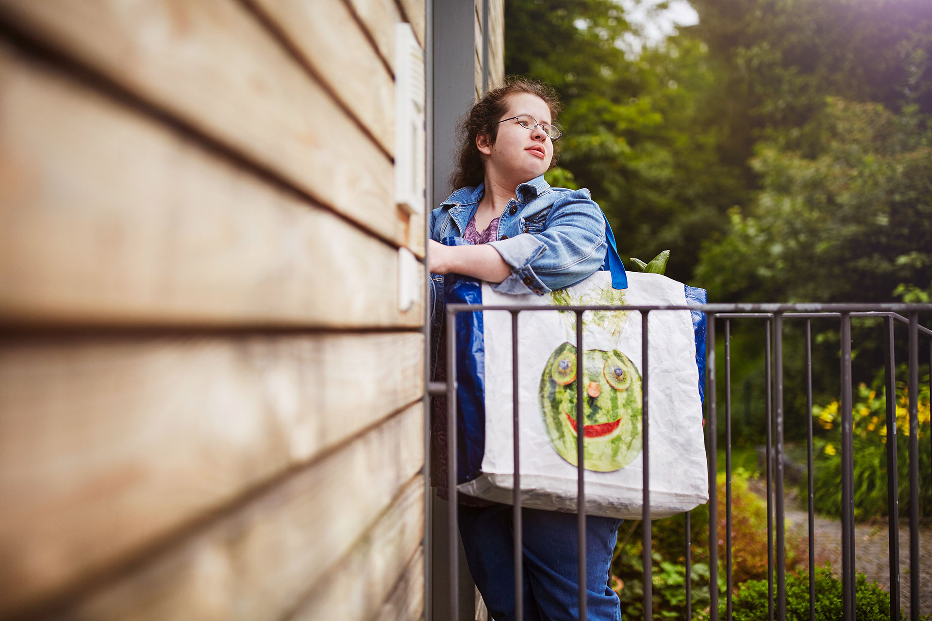 Im Bild: Eine Junge Frau steht an der Eingangstür eines Hauses und öffnet die Tür. Sie trägt eine große Tasche mit Lebensmitteln über der Schulter.