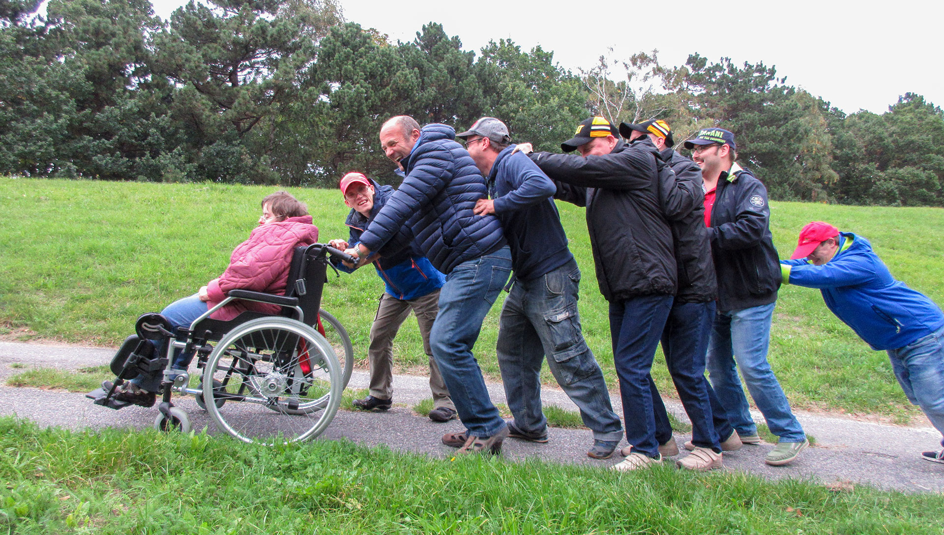 Ein Symbolfoto für Ehrenamt zeigt ein lustige Menschenkette, die eine Frau im Rollstuhl schiebt.