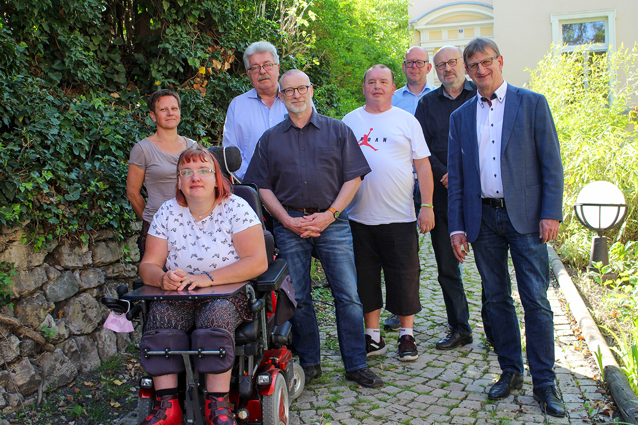 Gezeigt wird ein Gruppenfoto mit den 8 Mitgliedern des Lebenshilfe-Vorstandes Jena. Sie stehen bei sonnigem Wetter auf dem gepflasterten Weg vor dem Gebäuzde.