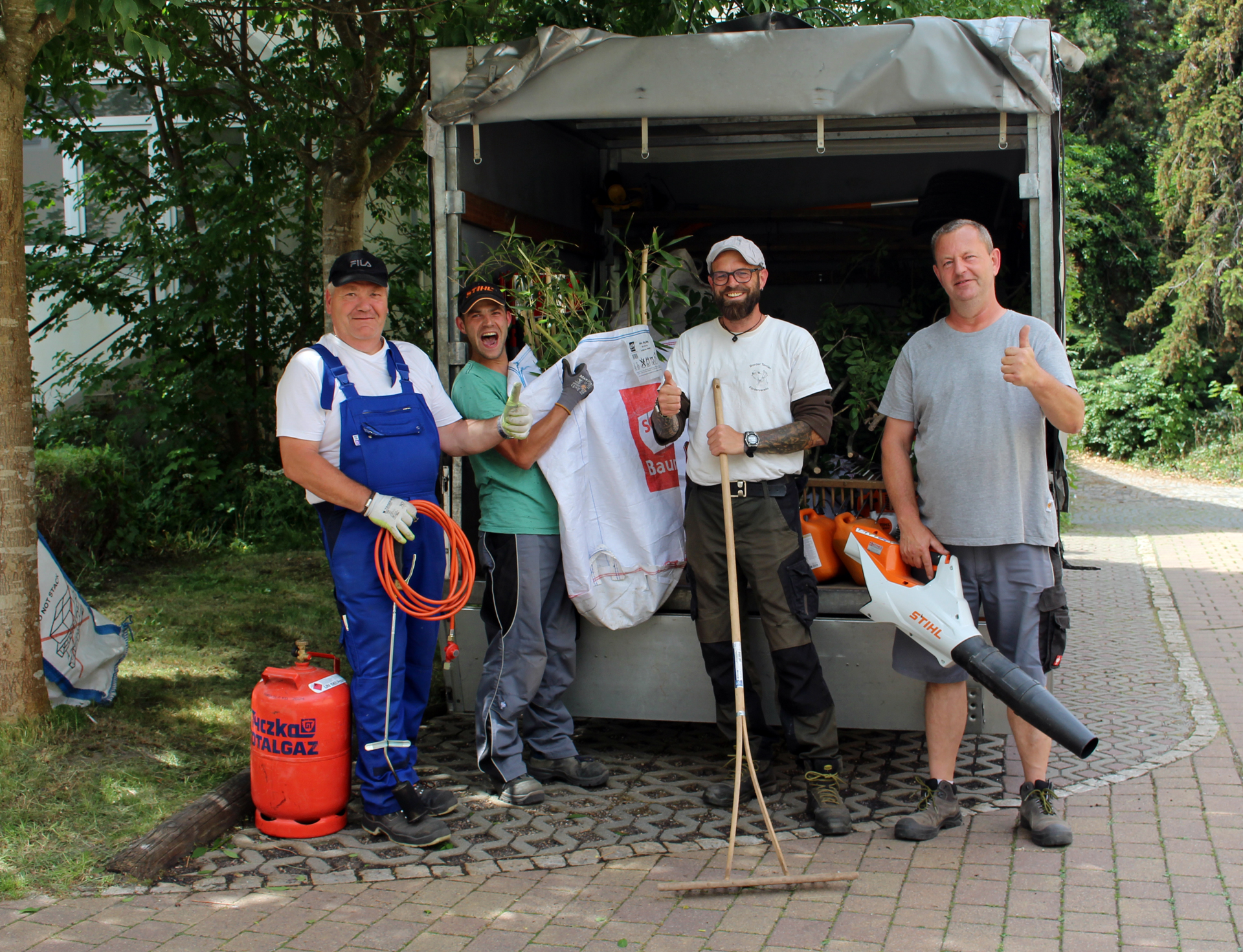Im Bild: Vier Mitarbeiter der SBW Service stehen fröhlich an der Ladefläche eines Transporters. Sie halten Werkzeuge aus dem Gartenbau in den Armen und zeigen den Daumen hoch.