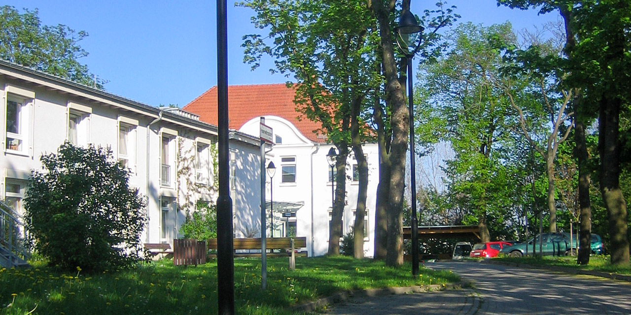 Abgebildet ist das weißes Wohnhaus mit begrünter Außernanlage und Zufahrt.